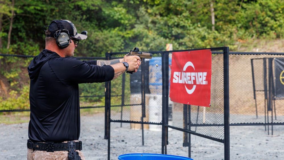 A shooter is actively firing a pistol at an outdoor shooting range, with a visible brass casing being ejected from the firearm. The range features a SureFire branded banner on a chain-link fence gate, with targets visible downrange. The setting appears to be a professional or competitive shooting event.