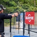 A shooter is actively firing a pistol at an outdoor shooting range, with a visible brass casing being ejected from the firearm. The range features a SureFire branded banner on a chain-link fence gate, with targets visible downrange. The setting appears to be a professional or competitive shooting event.