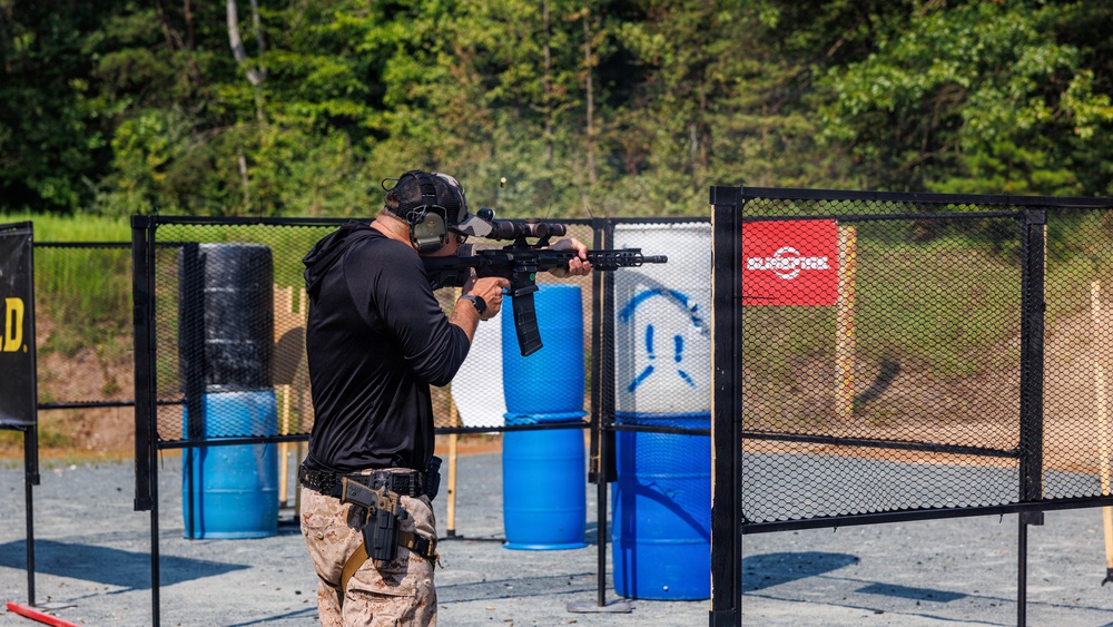 A shooter is participating in a competitive shooting event at an outdoor range, aiming a rifle equipped with a scope and extended magazine through a metal mesh gate. The shooter is wearing full protective gear including ear protection, a cap, and a holstered sidearm. The range features blue and red barrels as obstacles and paper targets in the background.