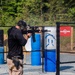 A shooter is participating in a competitive shooting event at an outdoor range, aiming a rifle equipped with a scope and extended magazine through a metal mesh gate. The shooter is wearing full protective gear including ear protection, a cap, and a holstered sidearm. The range features blue and red barrels as obstacles and paper targets in the background.