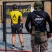 Two individuals are at an outdoor shooting range. The person in the foreground wears a black 'JMAP - Joint Marksmanship Assessment Package' hoodie and tactical gear, while the person in the background wears a yellow 'Apollo Arms' jersey and is actively shooting at targets. A Leupold-branded banner is visible in the background.