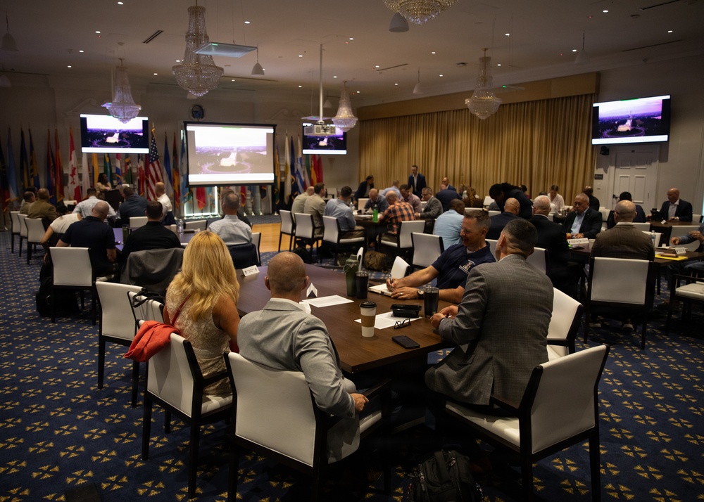 A formal conference or briefing session is taking place in an upscale ballroom-style venue decorated with international flags and crystal chandeliers. Attendees are seated at round and rectangular tables facing a presentation area with multiple screens displaying aerial imagery. The event appears to be a military or government-affiliated gathering based on the flags, attire mix of civilian and military dress, and professional setting.