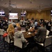 A formal conference or briefing session is taking place in an upscale ballroom-style venue decorated with international flags and crystal chandeliers. Attendees are seated at round and rectangular tables facing a presentation area with multiple screens displaying aerial imagery. The event appears to be a military or government-affiliated gathering based on the flags, attire mix of civilian and military dress, and professional setting.