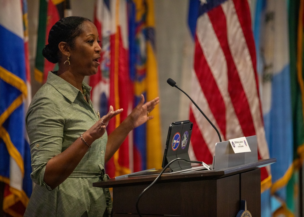 A woman in a green dress is speaking at a wooden podium in what appears to be an official or military setting. She is gesturing with both hands raised, suggesting an engaged and animated presentation. Multiple national and state flags are displayed in the background, along with an American flag.
