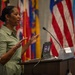 A woman in a green dress is speaking at a wooden podium in what appears to be an official or military setting. She is gesturing with both hands raised, suggesting an engaged and animated presentation. Multiple national and state flags are displayed in the background, along with an American flag.