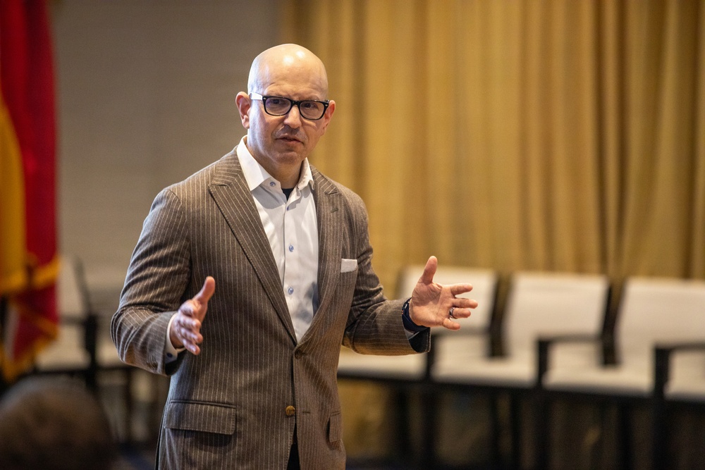 A bald man wearing glasses and a brown pinstripe blazer over a white dress shirt is speaking or presenting in what appears to be a conference or meeting room. He is gesturing with both hands in an expressive manner, suggesting active engagement with an audience. The background shows empty chairs, gold curtains, and what appears to be a flag on the left side.
