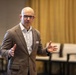 A bald man wearing glasses and a brown pinstripe blazer over a white dress shirt is speaking or presenting in what appears to be a conference or meeting room. He is gesturing with both hands in an expressive manner, suggesting active engagement with an audience. The background shows empty chairs, gold curtains, and what appears to be a flag on the left side.