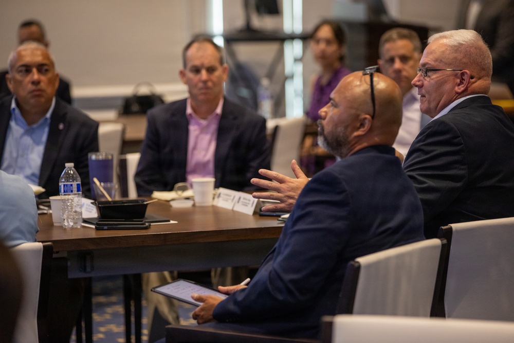 A formal meeting or roundtable discussion is taking place in what appears to be a conference or hotel meeting room. Several men in business attire are seated at tables, with one man in a navy suit gesturing with his hand while speaking to a man in a dark suit with glasses. Additional attendees are visible in the background observing the discussion.