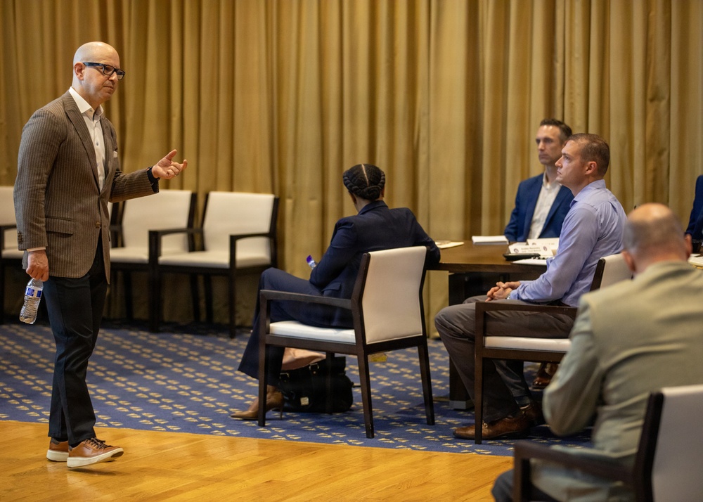 A bald man wearing glasses and a brown blazer appears to be presenting or speaking to a small group of seated attendees in what looks like a hotel conference or meeting room. He is gesturing with his right hand while holding a water bottle in his left, suggesting an active presentation or discussion. The audience of at least four people is seated at tables arranged in a semi-informal configuration.