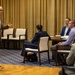 A bald man wearing glasses and a brown blazer appears to be presenting or speaking to a small group of seated attendees in what looks like a hotel conference or meeting room. He is gesturing with his right hand while holding a water bottle in his left, suggesting an active presentation or discussion. The audience of at least four people is seated at tables arranged in a semi-informal configuration.