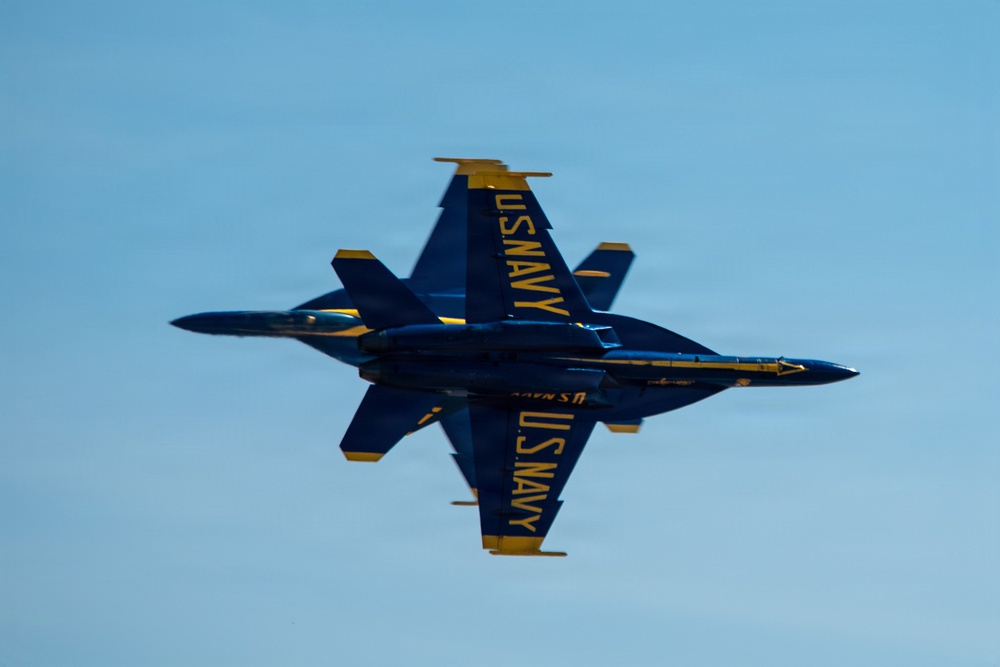 Two U.S. Navy Blue Angels F/A-18 Hornet jets are performing a close formation maneuver, appearing to fly in opposite vertical orientations directly on top of each other. The aircraft are painted in the iconic Blue Angels blue and gold color scheme with 'U.S. NAVY' lettering clearly visible on both fuselages. The image captures a dramatic belly-to-belly or mirror-image pass against a clear blue sky.