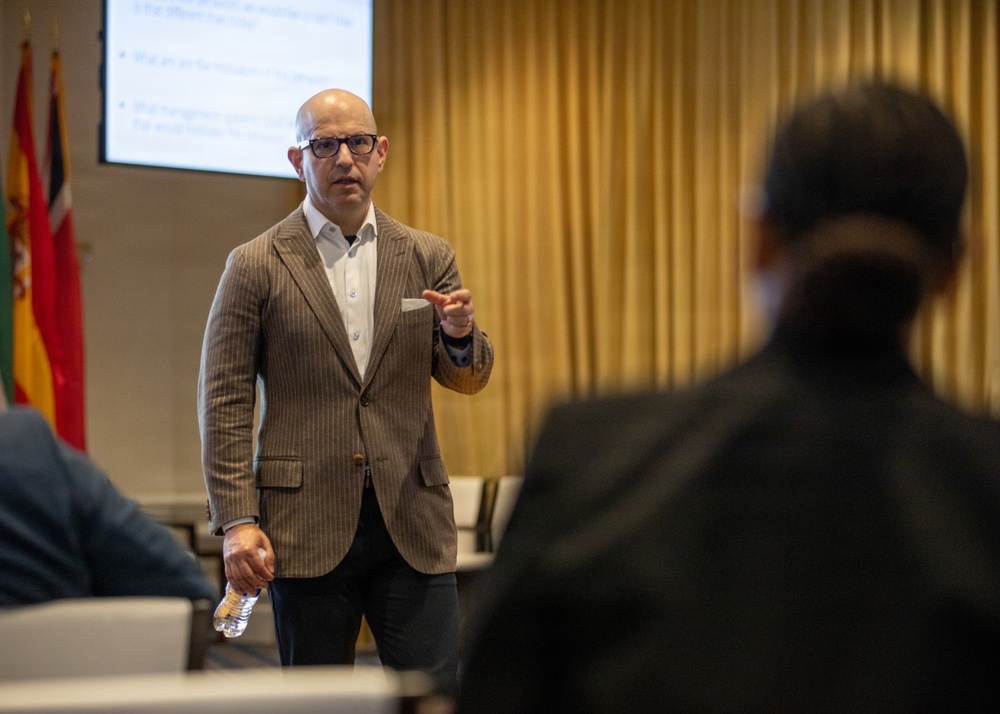 A bald man wearing glasses and a brown pinstripe blazer is delivering a presentation to an audience in a conference or meeting room. He is gesturing with one hand while holding a water bottle in the other, with a projected presentation slide visible behind him. Several flags are visible in the background, suggesting an international or formal organizational setting.