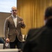 A bald man wearing glasses and a brown pinstripe blazer is delivering a presentation to an audience in a conference or meeting room. He is gesturing with one hand while holding a water bottle in the other, with a projected presentation slide visible behind him. Several flags are visible in the background, suggesting an international or formal organizational setting.