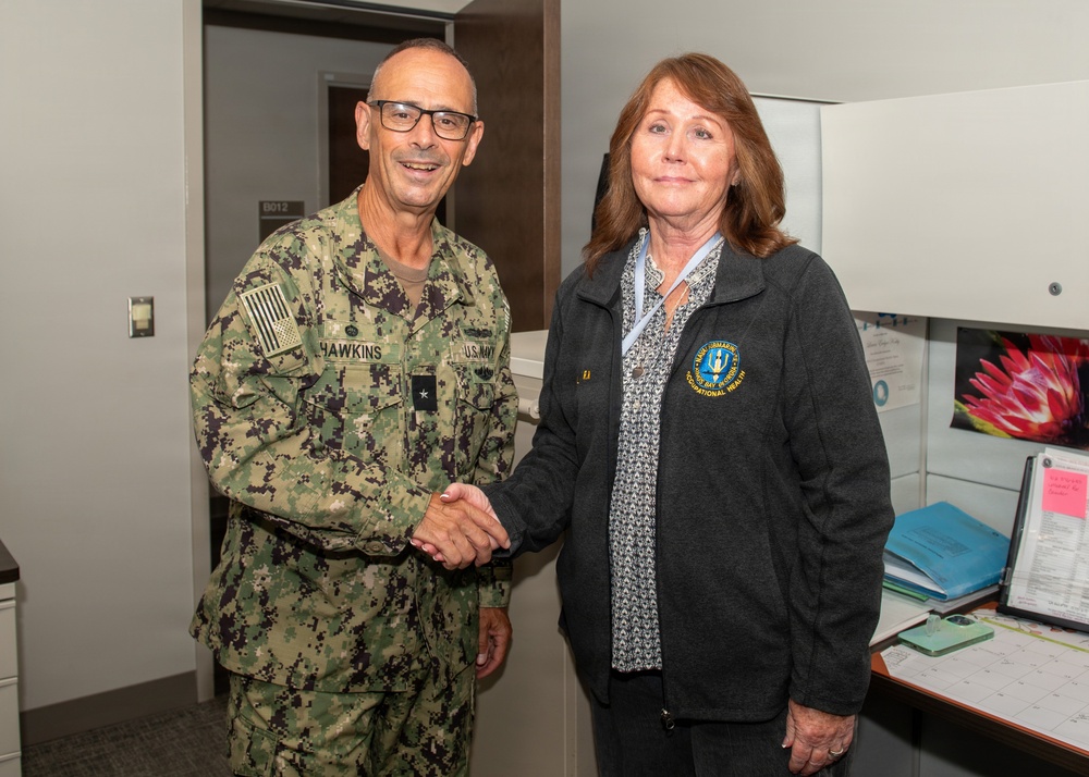 A U.S. Navy officer in digital camouflage uniform with the name tape 'HAWKINS' and a single star insignia (Rear Admiral lower half or Commodore) shakes hands with a civilian woman wearing a dark fleece jacket bearing an embroidered patch related to 'Occupational Health.' The setting appears to be an office or medical facility interior. The interaction appears formal and ceremonial in nature.