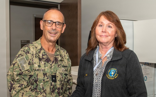 A U.S. Navy officer in digital camouflage uniform with the name tape 'HAWKINS' and a single star insignia (Rear Admiral lower half or Commodore) shakes hands with a civilian woman wearing a dark fleece jacket bearing an embroidered patch related to 'Occupational Health.' The setting appears to be an office or medical facility interior. The interaction appears formal and ceremonial in nature.