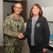 A U.S. Navy officer in digital camouflage uniform with the name tape 'HAWKINS' and a single star insignia (Rear Admiral lower half or Commodore) shakes hands with a civilian woman wearing a dark fleece jacket bearing an embroidered patch related to 'Occupational Health.' The setting appears to be an office or medical facility interior. The interaction appears formal and ceremonial in nature.