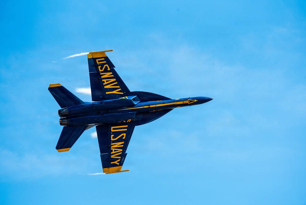 A U.S. Navy Blue Angels F/A-18 Hornet jet is captured in an inverted flight maneuver against a clear blue sky. The aircraft displays the iconic Blue Angels livery of deep navy blue with bold yellow 'U.S. NAVY' lettering visible on both the top and bottom fuselage. Vapor trails are visible streaming from the wingtips, indicating high-speed, high-G flight conditions.