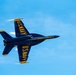 A U.S. Navy Blue Angels F/A-18 Hornet jet is captured in an inverted flight maneuver against a clear blue sky. The aircraft displays the iconic Blue Angels livery of deep navy blue with bold yellow 'U.S. NAVY' lettering visible on both the top and bottom fuselage. Vapor trails are visible streaming from the wingtips, indicating high-speed, high-G flight conditions.