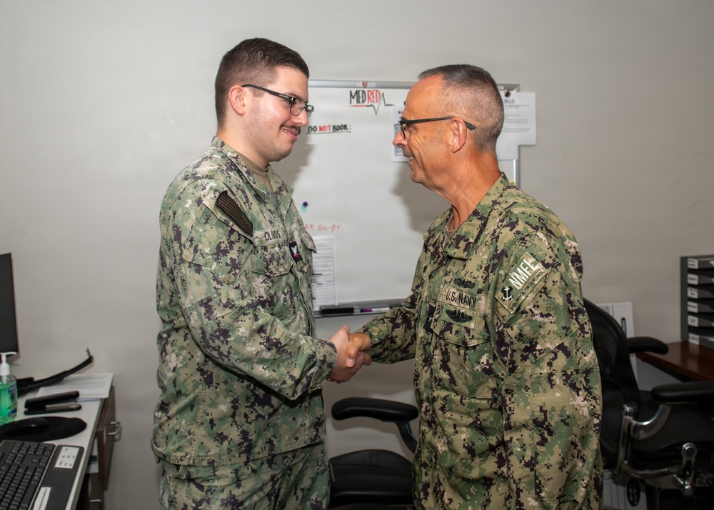 Two U.S. Navy personnel in NWU Type III (green digital camouflage) uniforms are shaking hands in what appears to be an office or administrative space. The individual on the right displays a 'U.S. Navy' tape and an anchor insignia indicating senior enlisted or officer rank, while the individual on the left has a name tape reading 'OLIVOS'. The setting appears to be a formal recognition or greeting moment within a military facility.