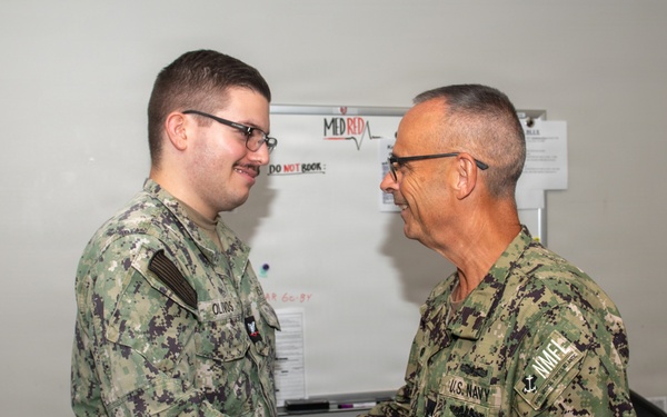Two U.S. Navy personnel in NWU Type III (green digital camouflage) uniforms are shaking hands in what appears to be an office or administrative space. The individual on the right displays a 'U.S. Navy' tape and an anchor insignia indicating senior enlisted or officer rank, while the individual on the left has a name tape reading 'OLIVOS'. The setting appears to be a formal recognition or greeting moment within a military facility.