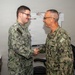 Two U.S. Navy personnel in NWU Type III (green digital camouflage) uniforms are shaking hands in what appears to be an office or administrative space. The individual on the right displays a 'U.S. Navy' tape and an anchor insignia indicating senior enlisted or officer rank, while the individual on the left has a name tape reading 'OLIVOS'. The setting appears to be a formal recognition or greeting moment within a military facility.