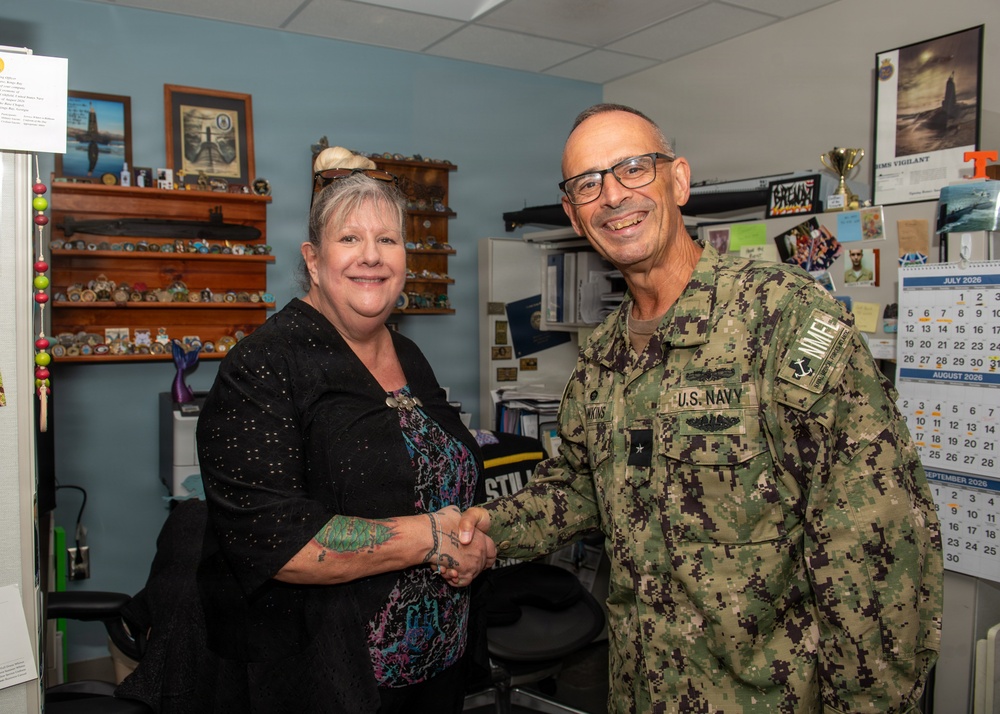 A U.S. Navy officer in NWU Type III (digital camouflage) uniform is shaking hands with a civilian woman in an office setting. The officer wears rank insignia consistent with a flag officer (one star visible) and submarine warfare qualification badges. The setting appears to be a military administrative office decorated with naval memorabilia, challenge coins, and submarine-related items.