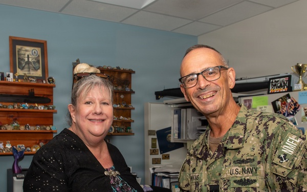 A U.S. Navy officer in NWU Type III (digital camouflage) uniform is shaking hands with a civilian woman in an office setting. The officer wears rank insignia consistent with a flag officer (one star visible) and submarine warfare qualification badges. The setting appears to be a military administrative office decorated with naval memorabilia, challenge coins, and submarine-related items.