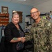 A U.S. Navy officer in NWU Type III (digital camouflage) uniform is shaking hands with a civilian woman in an office setting. The officer wears rank insignia consistent with a flag officer (one star visible) and submarine warfare qualification badges. The setting appears to be a military administrative office decorated with naval memorabilia, challenge coins, and submarine-related items.