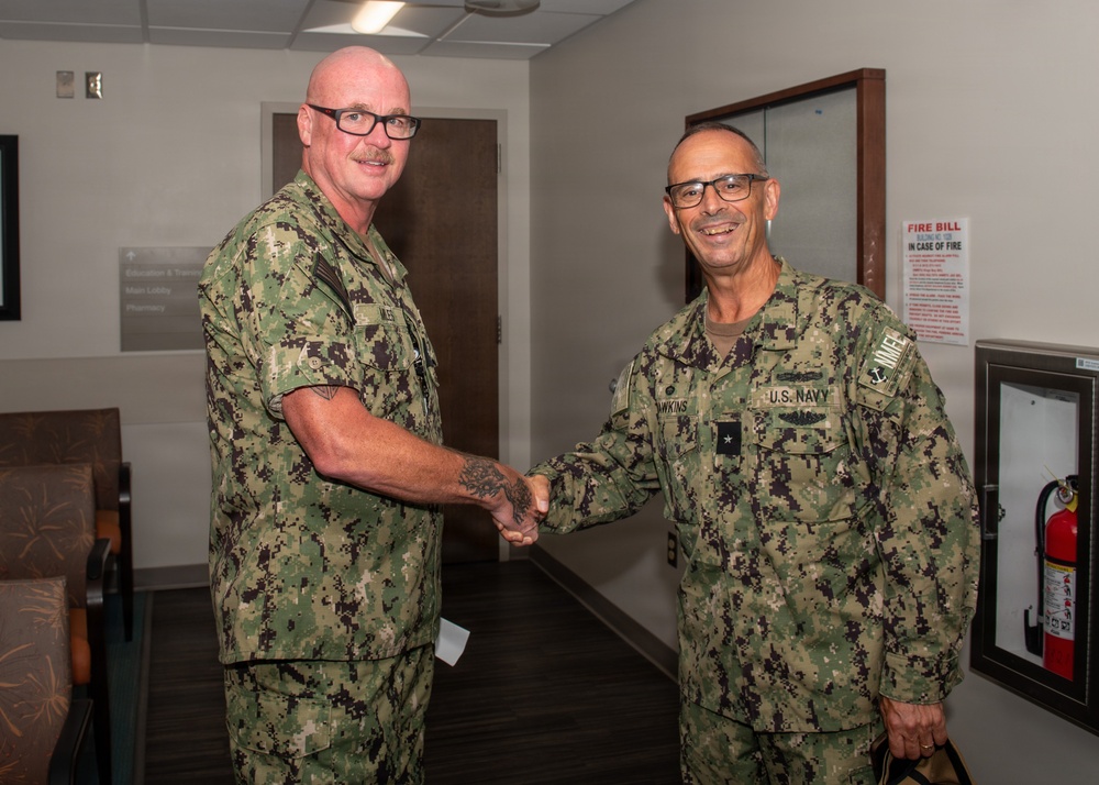 Two U.S. Navy personnel in Navy Working Uniform (NWU Type III) camouflage are shaking hands in what appears to be a military medical facility. The individual on the right has visible rank insignia and a name tape reading 'HAWKINS' with 'U.S. NAVY' tape, along with what appears to be a one-star flag officer rank. The setting includes directional signage for a lobby, pharmacy, and education areas, consistent with a naval medical center.