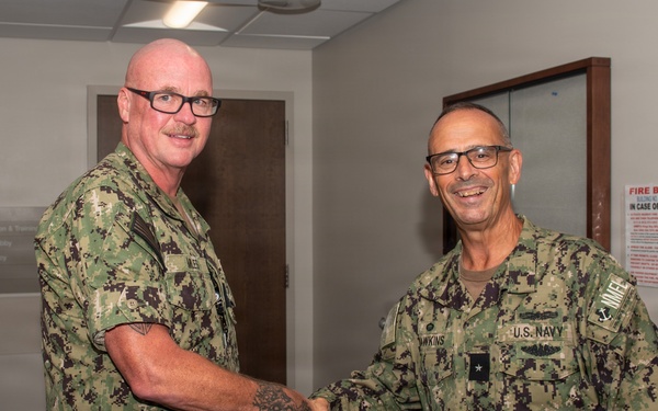 Two U.S. Navy personnel in Navy Working Uniform (NWU Type III) camouflage are shaking hands in what appears to be a military medical facility. The individual on the right has visible rank insignia and a name tape reading 'HAWKINS' with 'U.S. NAVY' tape, along with what appears to be a one-star flag officer rank. The setting includes directional signage for a lobby, pharmacy, and education areas, consistent with a naval medical center.
