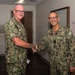Two U.S. Navy personnel in Navy Working Uniform (NWU Type III) camouflage are shaking hands in what appears to be a military medical facility. The individual on the right has visible rank insignia and a name tape reading 'HAWKINS' with 'U.S. NAVY' tape, along with what appears to be a one-star flag officer rank. The setting includes directional signage for a lobby, pharmacy, and education areas, consistent with a naval medical center.