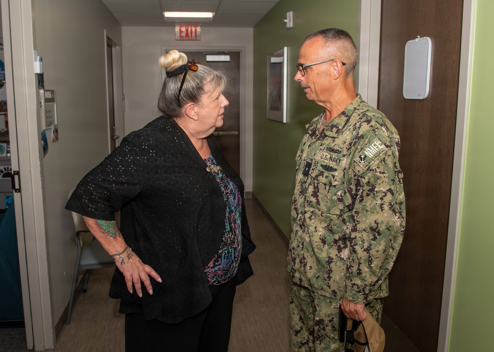 A civilian woman and a U.S. Navy servicemember are engaged in conversation in what appears to be an indoor hallway or office corridor. The Navy personnel is wearing the standard Navy Working Uniform (NWU) camouflage pattern with visible 'U.S. NAVY' tape and insignia. The setting appears to be a military or government facility based on the institutional interior design.