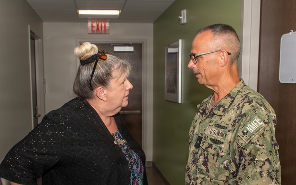 A civilian woman and a U.S. Navy servicemember are engaged in conversation in what appears to be an indoor hallway or office corridor. The Navy personnel is wearing the standard Navy Working Uniform (NWU) camouflage pattern with visible 'U.S. NAVY' tape and insignia. The setting appears to be a military or government facility based on the institutional interior design.