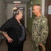 A civilian woman and a U.S. Navy servicemember are engaged in conversation in what appears to be an indoor hallway or office corridor. The Navy personnel is wearing the standard Navy Working Uniform (NWU) camouflage pattern with visible 'U.S. NAVY' tape and insignia. The setting appears to be a military or government facility based on the institutional interior design.