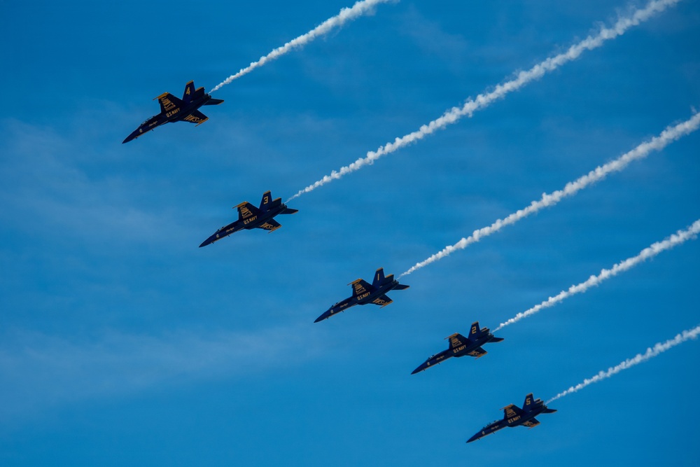 Five U.S. Navy Blue Angels F/A-18 Hornet jets fly in a diagonal echelon formation against a clear blue sky. Each aircraft is numbered (1 through 5, visible on the fuselages) and emits white smoke trails. The image captures a precision aerobatic demonstration with sharp detail and vivid color contrast.