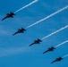 Five U.S. Navy Blue Angels F/A-18 Hornet jets fly in a diagonal echelon formation against a clear blue sky. Each aircraft is numbered (1 through 5, visible on the fuselages) and emits white smoke trails. The image captures a precision aerobatic demonstration with sharp detail and vivid color contrast.