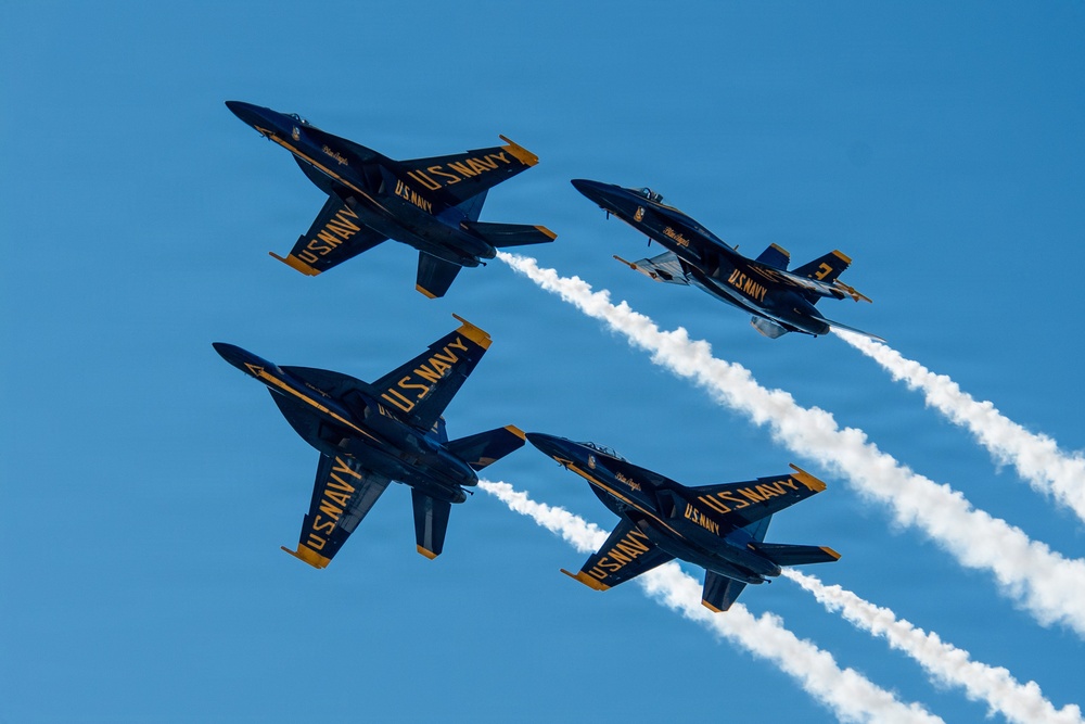 Four U.S. Navy Blue Angels F/A-18 Hornet jets are performing a formation flight maneuver against a clear blue sky. The aircraft are flying in a tight diamond-like formation, with white smoke trails visible from at least three of the jets. The markings 'U.S. Navy' and 'Blue Angels' are clearly legible on the fuselages, with distinctive navy blue and gold color schemes.