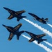 Four U.S. Navy Blue Angels F/A-18 Hornet jets are performing a formation flight maneuver against a clear blue sky. The aircraft are flying in a tight diamond-like formation, with white smoke trails visible from at least three of the jets. The markings 'U.S. Navy' and 'Blue Angels' are clearly legible on the fuselages, with distinctive navy blue and gold color schemes.