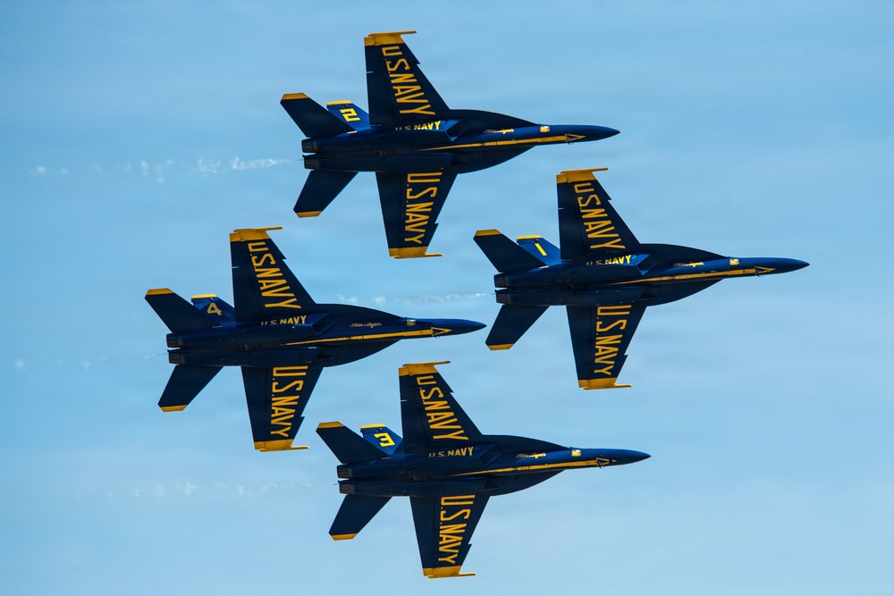 Four U.S. Navy Blue Angels aircraft are flying in a tight diamond formation against a clear blue sky. The jets are F/A-18 Hornets painted in the iconic blue and gold color scheme with 'U.S. NAVY' markings. The aircraft are numbered 1 through 4, representing the signature diamond formation used in Blue Angels air show performances.