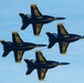 Four U.S. Navy Blue Angels aircraft are flying in a tight diamond formation against a clear blue sky. The jets are F/A-18 Hornets painted in the iconic blue and gold color scheme with 'U.S. NAVY' markings. The aircraft are numbered 1 through 4, representing the signature diamond formation used in Blue Angels air show performances.