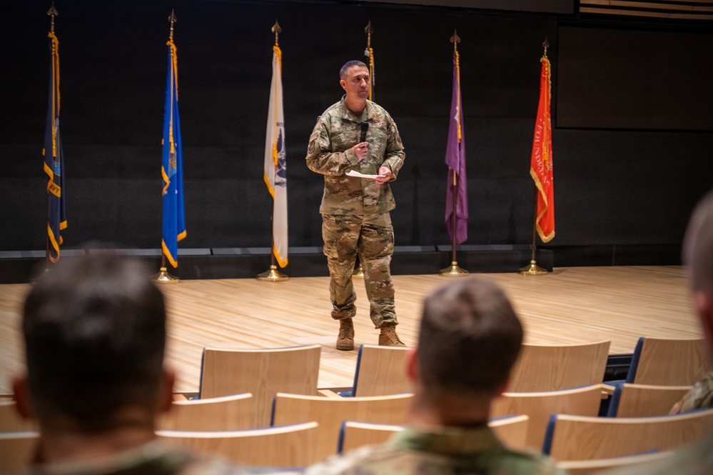 A military officer in OCP (Operational Camouflage Pattern) uniform stands on a stage addressing an audience of seated military personnel in an auditorium setting. Multiple military unit flags and guidons are displayed on stands behind the speaker. The setting appears to be a formal military briefing, ceremony, or command presentation.
