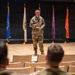 A military officer in OCP (Operational Camouflage Pattern) uniform stands on a stage addressing an audience of seated military personnel in an auditorium setting. Multiple military unit flags and guidons are displayed on stands behind the speaker. The setting appears to be a formal military briefing, ceremony, or command presentation.