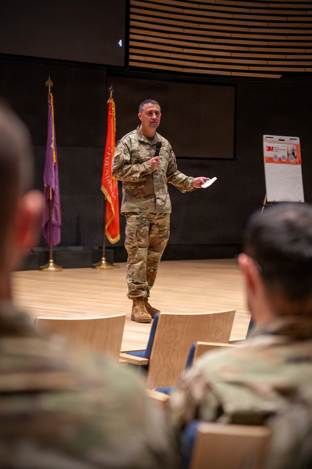 A U.S. Army soldier in Operational Camouflage Pattern (OCP) uniform is delivering a presentation or briefing on a stage to a seated military audience. The speaker is holding a microphone and what appears to be note cards or a small device. The setting appears to be a formal auditorium or briefing room with military flags displayed on stage.