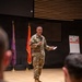 A U.S. Army soldier in Operational Camouflage Pattern (OCP) uniform is delivering a presentation or briefing on a stage to a seated military audience. The speaker is holding a microphone and what appears to be note cards or a small device. The setting appears to be a formal auditorium or briefing room with military flags displayed on stage.