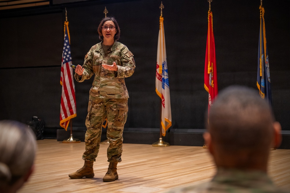 A female U.S. military service member in OCP (Operational Camouflage Pattern) uniform is addressing an audience from a stage or platform. She appears to be giving a briefing or presentation, gesturing with her hands while speaking. Several military and organizational flags are displayed behind her, and audience members are visible in the foreground.