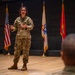 A female U.S. military service member in OCP (Operational Camouflage Pattern) uniform is addressing an audience from a stage or platform. She appears to be giving a briefing or presentation, gesturing with her hands while speaking. Several military and organizational flags are displayed behind her, and audience members are visible in the foreground.