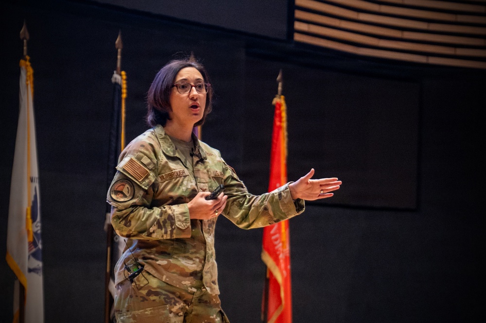 A U.S. Army soldier with the name tape 'YARBROUGH' is delivering a presentation or briefing in what appears to be an auditorium or conference hall. She is wearing Operational Camouflage Pattern (OCP) uniform with a U.S. flag patch on the right sleeve and holds a presentation clicker. Military flags and guidons are visible in the background.
