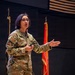 A U.S. Army soldier with the name tape 'YARBROUGH' is delivering a presentation or briefing in what appears to be an auditorium or conference hall. She is wearing Operational Camouflage Pattern (OCP) uniform with a U.S. flag patch on the right sleeve and holds a presentation clicker. Military flags and guidons are visible in the background.