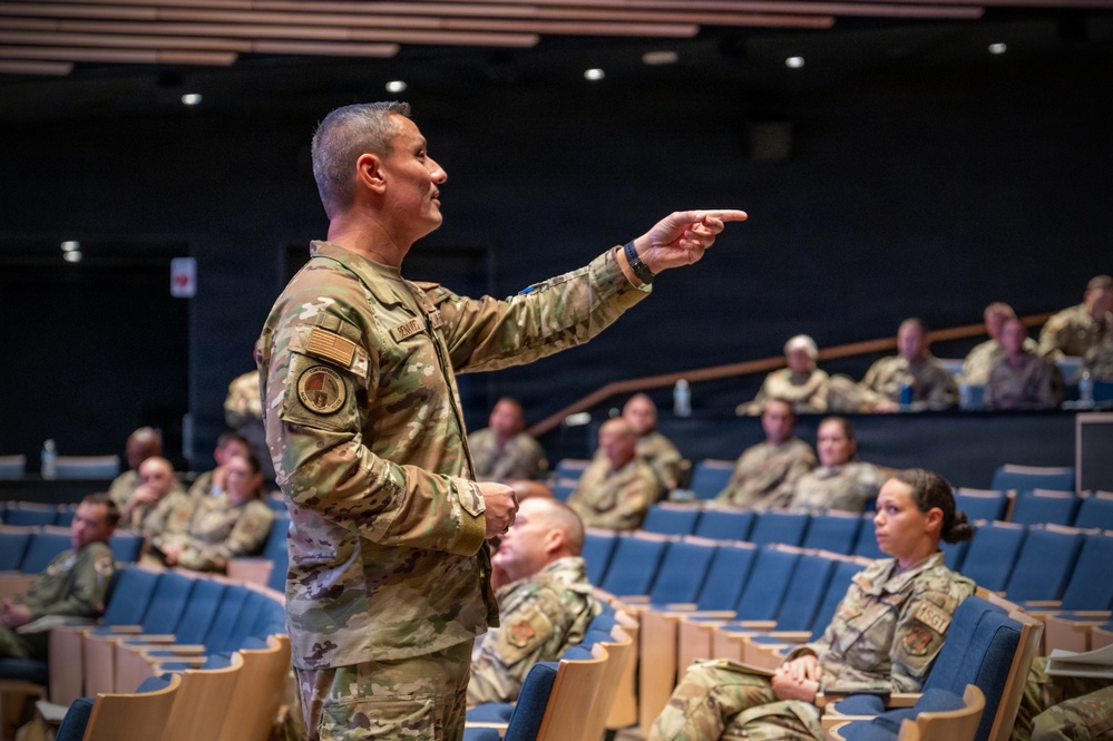 A military officer in OCP (Operational Camouflage Pattern) uniform is addressing an audience of fellow service members in a modern auditorium. The speaker is pointing toward the audience while delivering what appears to be a briefing or training session. The audience consists of multiple uniformed military personnel seated in blue auditorium chairs.