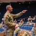 A military officer in OCP (Operational Camouflage Pattern) uniform is addressing an audience of fellow service members in a modern auditorium. The speaker is pointing toward the audience while delivering what appears to be a briefing or training session. The audience consists of multiple uniformed military personnel seated in blue auditorium chairs.